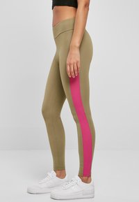 Olivengrønne leggings med en rosa sideStripe, laget av stretchstoff. De har høy midje og er parret med hvite sneakers.