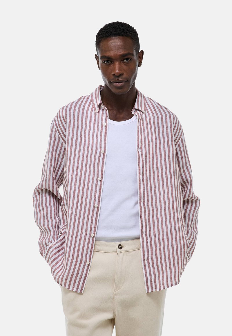 Chemise à rayures à manches longues en blanc et rouge clair, fabriquée en tissu avec un col boutonné. Portée ouverte sur un débardeur blanc.