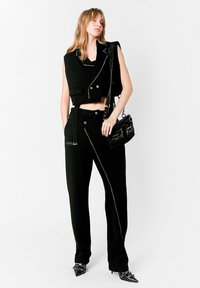 Chaleco corto negro con detalles plateados, pantalones de sastre con cremallera asimétrica, combinado con un bolso pequeño negro y tacones puntiagudos.