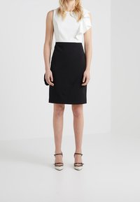 Haut sans manches blanc avec des volants, associé à une jupe noire près du corps avec des accents de couture diagonaux. Le mannequin porte des chaussures à talons noir et blanc.