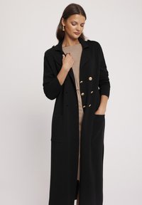 Czarny długi płaszcz z złotymi guzikami, z wyłogami, dwoma przednimi kieszeniami, strukturalnym designem i prążkowaną teksturą. Nosi się go na beżowej sukience.