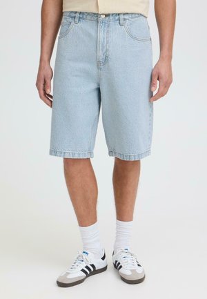 Homme portant un short en jean bleu clair, des chaussettes blanches et des baskets blanches à rayures noires, debout devant un fond uni.