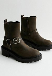 Bruna ankelstövlar med strukturerad yta, sidoglittring, spännedetalj och tjock svart sula. Har en robust och tuff design.