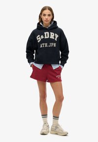 Marine hoodie met "S&D" graphics, gecombineerd met bordeauxrode shorts met geborduurde "SUPERDRY NYC." Afgemaakt met grijze sneakers en gestreepte sokken.
