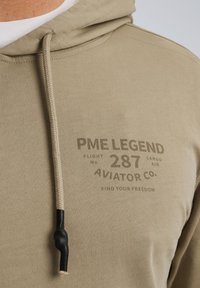 Sweat à capuche khaki en tissu doux, affichant un imprimé sur le devant avec "PME LEGEND", et un cordon de serrage avec un embout en caoutchouc noir.