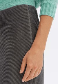 Grauer Cordrock mit gerippter Textur, ausgestattet mit einem seitlichen Reißverschluss und einer eng anliegenden Silhouette. Eine Hand ruht sanft auf dem Stoff.