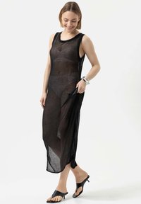 Schwarzes, durchsichtiges Strickkleid mit ärmellosem Design, seitlichem Schlitz und rundem Ausschnitt. Über einem schwarzen Unterkleid getragen, ergänzt durch schwarze Sandalen.