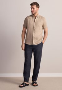 Beige Leinenhemd mit kurzen Ärmeln und einer Tasche, kombiniert mit dunkelblauen Leinenhosen und schwarzen Slide-Sandalen. Schlichtes, locker sitzendes Design.