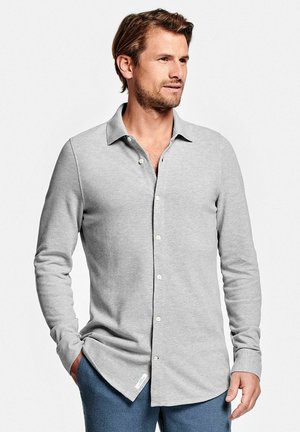 Homme aux cheveux châtain clair et barbe, portant une chemise grise claire à manches longues boutonnée et un pantalon bleu, debout avec une main dans la poche.