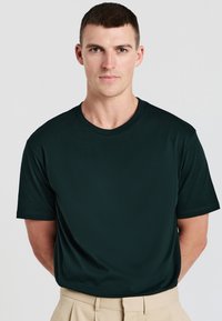 Giovane uomo con i capelli corti che indossa una maglietta semplice a girocollo verde scuro e pantaloni beige, in piedi contro uno sfondo chiaro.