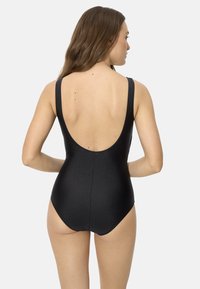 Mujer con un bañador de una pieza negro con espalda baja en forma de escote redondo, de pie con las manos en las caderas sobre un fondo liso.