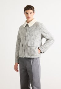 Tânăr îmbrăcat cu o jachetă gri în carouri, cu fermoar și guler din fleece alb, purtând pantaloni gri, stând cu o mână în buzunar.