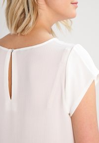 Blusa bianca con maniche corte, caratterizzata da un'apertura a occhiello nella parte posteriore, realizzata in tessuto leggero e testurizzato.