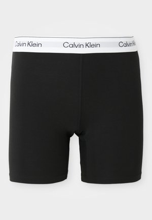 Crne bokserice s prilagođenim dizajnom, rastezljivim materijalom i bijelom Calvin Klein logom na pojasu. Glatka tekstura i dužina do sredine bedara.