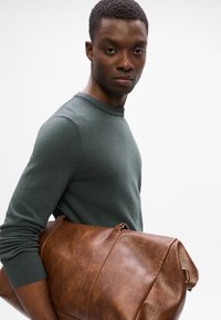 Brun læder duffelbag med en tekstureret overflade og sølvhardware, holdt under armen af en person iført en mørkegrøn langærmet skjorte.