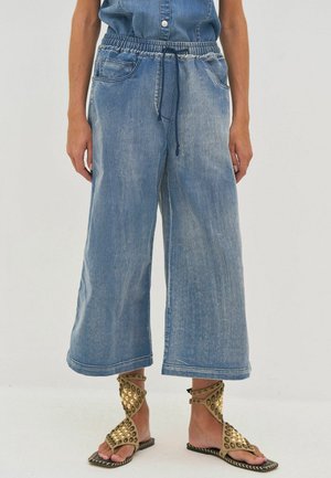 Személy széles szárú, rövidített farmernadrágot visel, rugalmas derékkal és sötétkék zsinórral, bézs színű, szegecses, bokánál megkötős szandállal párosítva.