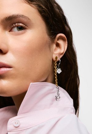 Primer plano de una persona que lleva un pendiente de cadena dorada con un charm plateado "Tauri" y un pequeño colgante cuadrado plateado, vestido con una camisa de cuello rosa claro.