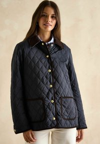 Pikowana granatowa kurtka z brązowym kołnierzem z sztruksu, złotymi elementami wykończenia oraz dwiema przednimi kieszeniami z brązowym obszyciem i wyszytym logo.