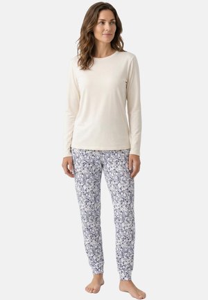 Kobieta ubrana w białą koszulę z długim rękawem oraz białe spodnie do spania z niebieskim kwiatowym wzorem, stojąca boso na jednolitym tle.