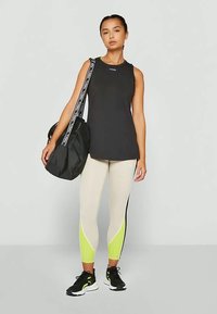 Crni top bez rukava, bež tajice s žutim naglasima i crne atletske cipele. Model drži crnu torbu s mrežastim panelom.