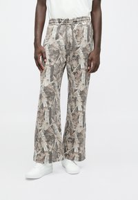 Levélmintás, földszínekben tündöklő, elastikus derekú nadrág, széles lábakkal és laza szabással, fehér sportcipőkkel párosítva.