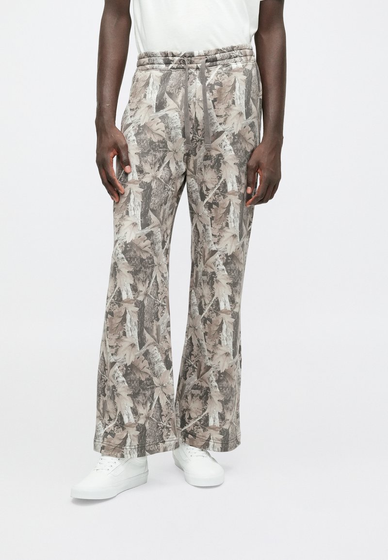 Levélmintás, földszínekben tündöklő, elastikus derekú nadrág, széles lábakkal és laza szabással, fehér sportcipőkkel párosítva.