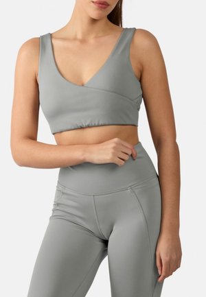 Vrouw draagt lichtgrijze cropped sportbeha en hooggetailleerde legging, staat met één gebogen arm en hand dicht bij de taille.