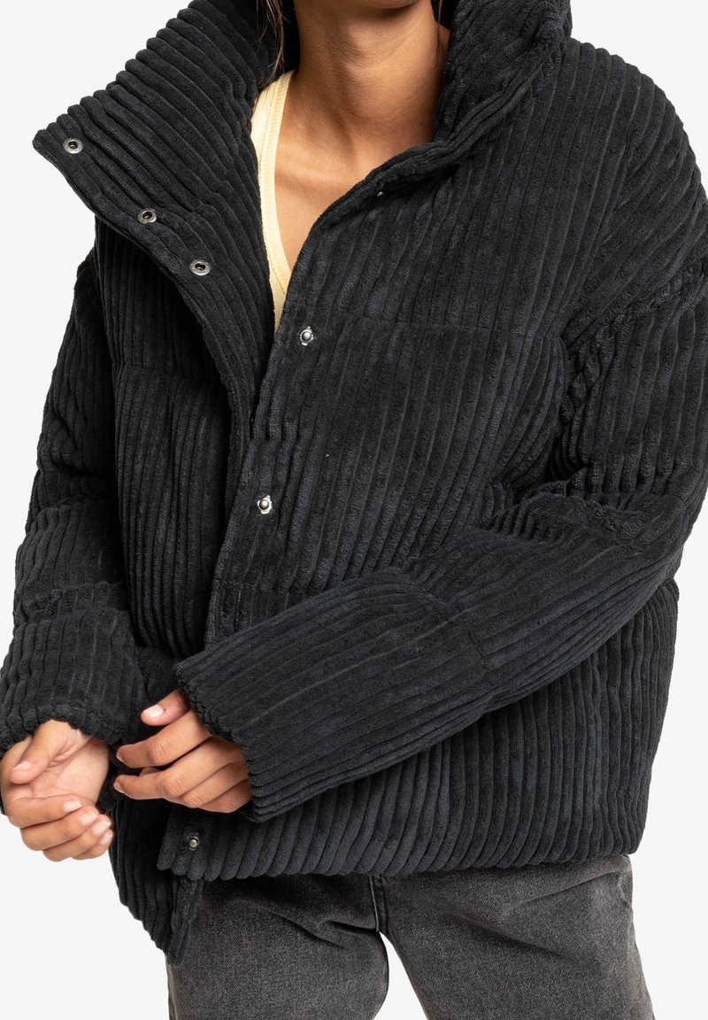 Personne portant une veste matelassée en velours côtelé noir à boutons pression, chemise beige en dessous et jean foncé délavé, mains partiellement jointes.