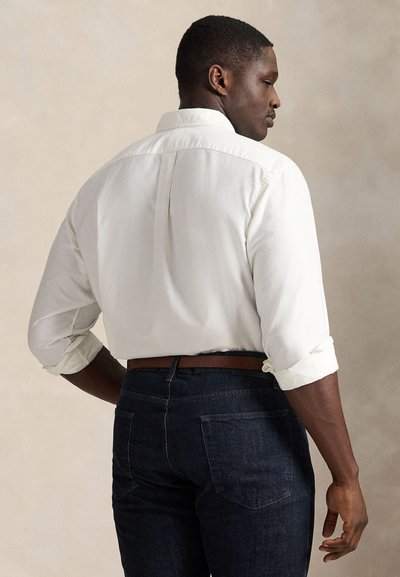 Mand iført en hvid langærmet skjorte med opkærmede ærmer og mørkeblå jeans, stående sidevej mod en ensfarvet baggrund.