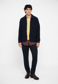 Veste en mélange de laine marine avec deux poches avant, superposée sur un pull en maille jaune et une chemise à motifs, associée à un pantalon foncé et des bottes brunes.