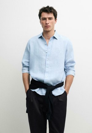 Junger Mann, der ein hellblaues Hemd mit hochgekrempelten Ärmeln trägt und einen schwarzen Pullover um die Taille gebunden hat, die Hände in den Taschen.