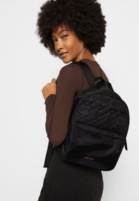 Mochila negra de nailon acolchado con respaldo acolchado, bolsillo frontal con cremallera y logotipo en dorado. Correas ajustables y una forma elegante y redondeada.