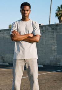 Ung mann står med armene i kors på en solfylt utendørs parkeringsplass, iført en beige t-skjorte og lyse bukser.