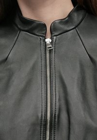 Casaco de couro preto com gola subida, fecho frontal com fecho éclair e ferragens prateadas, detalhes de costura subtil. Textura suave, design ajustado.