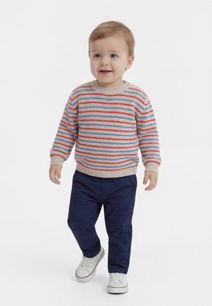 Maluch ubrany w beżowy sweter w paski, granatowe spodnie i białe trampki, idący do przodu na białym tle.