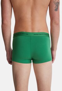 Torso masculino visto desde atrás, vistiendo boxers verdes con una cinturilla de "United Colors of Benetton", y un tatuaje de un ángel en el brazo izquierdo.