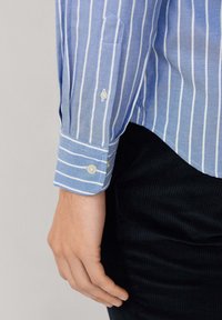 Camisa a rayas azul claro con líneas verticales blancas, con puños de botones y una tela suave y texturada. La mano descansa sobre pantalones de pana oscura.