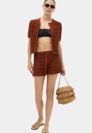 Kvinde iført brune strikkede shorts og kortærmet cardigan med knapper, leopardprint bandeau, bærende en frynset stråtaske, beige sandaler og solbriller.
