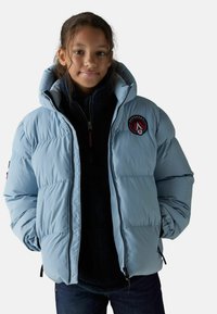 Doudoune bleu clair avec un col montant, présentant un logo rond sur la poitrine, des poches zippées et une doublure en peluche noire en dessous.