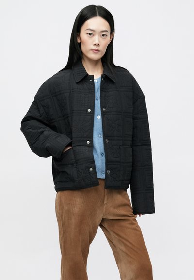 Mustriga musta jakki, millel on kastilise disain, tekstuuriga mustrid, nööpidega kinnihoidmine ja krae, kantud helesinise särgi ja pruunide pükste kohal.