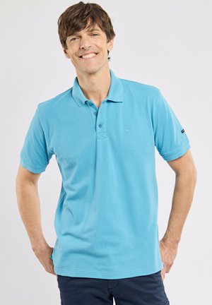 Homme souriant portant un polo bleu clair à manches courtes et un pantalon foncé, debout les mains sur les hanches devant un fond uni.