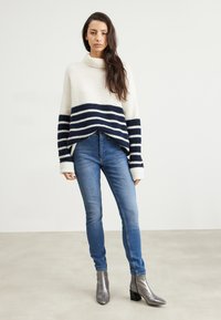 Suéter grueso de crema con rayas marinas, cuello alto y un corte corto, combinado con jeans azules ajustados y botines de tacón bloque metálico.