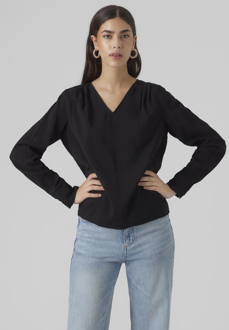 Maglietta nera a maniche lunghe con scollatura a V, dettagli arricciati sulle spalle, vestibilità comoda e tessuto testurizzato, abbinata a jeans azzurri chiari.