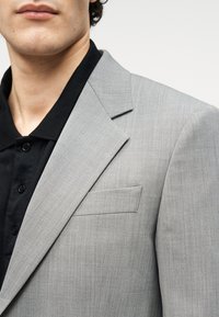 Ανοιχτό γκρι σακάκι από υφασμάτινη υφή, με γιακά με σχισμή και μια μονή τσέπη στο στήθος, συνδυασμένο με μαύρο πόλο μπλουζάκι από κάτω.