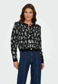 Femme portant un cardigan boutonné noir et gris à motifs, associé à un jean bleu taille haute, se tenant devant un fond uni.