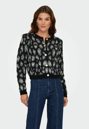 Vrouw in een zwart-grijs gemêleerde geknoopte cardigan, gecombineerd met hoge taille blauwe jeans, staand tegen een effen achtergrond.