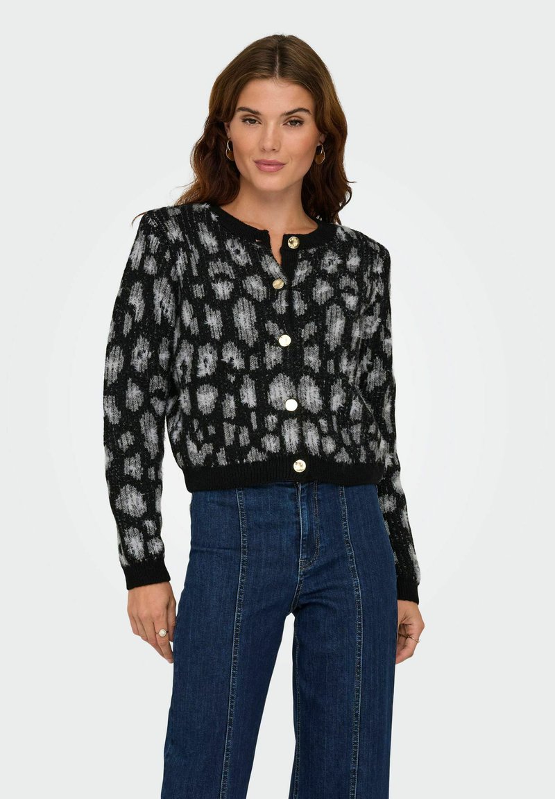 Femme portant un cardigan boutonné noir et gris à motifs, associé à un jean bleu taille haute, se tenant devant un fond uni.