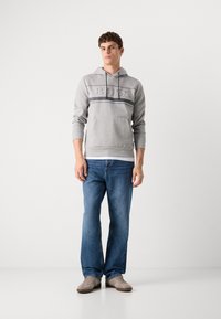 Gray hoodie with a textured "BOSS" logo and dark stripes, paired with blue denim jeans and beige sneakers. Model stands with a neutral pose.
