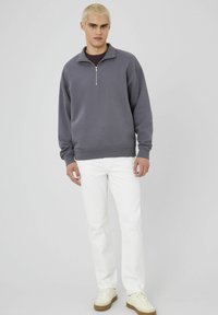 Sweatshirt zippé gris à manches longues, porté sur une chemise sombre, associé à un jean blanc et des baskets beiges. Couleur unie, design décontracté.