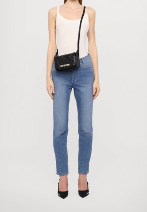 Schwarze, gesteppten Umhängetasche mit goldenem Logo, getragen mit einem figurbetonten weißen Tanktop und hellblauen, hochtaillierten Jeans, ergänzt durch schwarze Absätze.